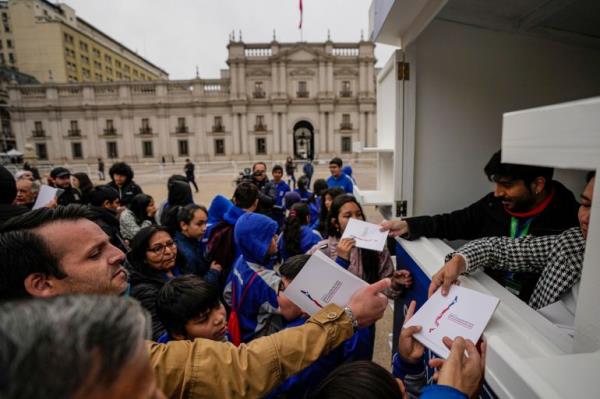 A crowd of people gather in front of the window of a booth in Santiago, wher<em></em>e people hand out copies of the latest co<em></em>nstitutional draft. The presidential palace is visible in the background.