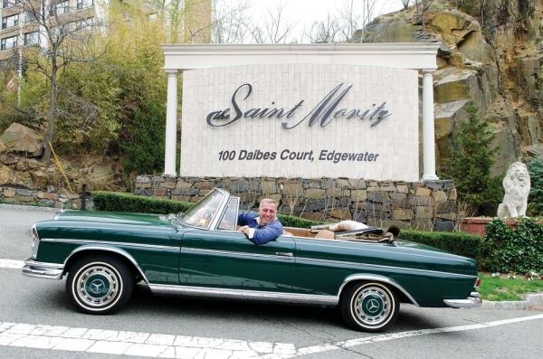 Fred Daibes at the wheel of an open top vintage Mercedes car outside a sign saying 