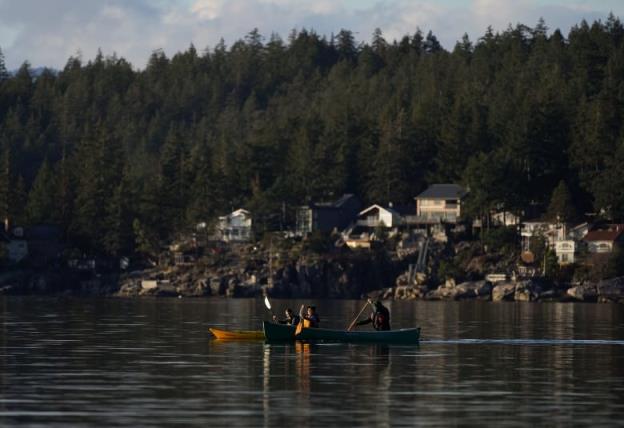 Two people kayak past a waterside village in the shadow of mountains.