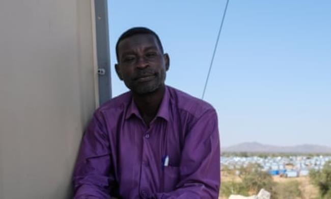 Gamar al-Deen, a teacher in El Geneina, wearing a purple shirt with a backdrop of buildings and distant mountains behind him