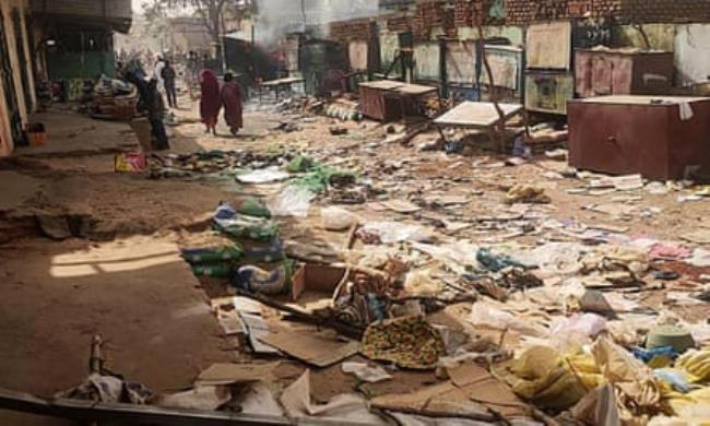 People walk along a street scattered with broken boxes and destroyed market stalls in El Geneina