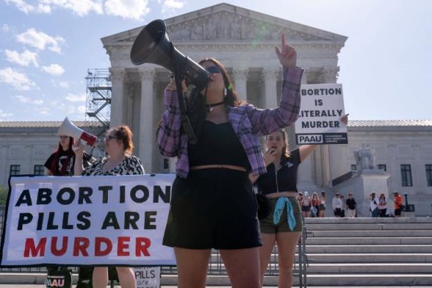 A woman wearing sunglasses, a flannel shirt and shorts is shown speaking into a megaphone at a demo<em></em>nstration in front of the steps leading to an august building. Near her, a sign reads, 'Abortion pills are murder.'