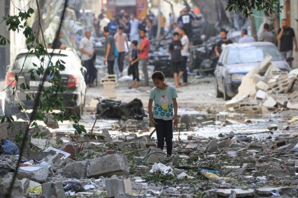 A Palestinian girl walks through the debris a day after the Israeli raid. There are damaged cars and men standing around in the background.