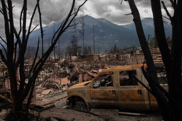 Mountains overlook a landscape filled with burned out rubble. A charred yellow truck sits in the foreground.