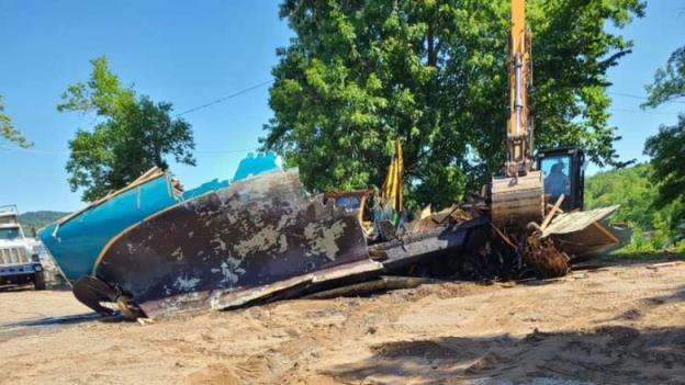 Two excavators dig into a large, battered sailboat on the beach, with a dump truck in the background.