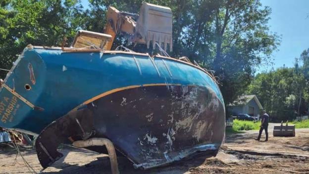 An excavator digs into a large, battered sailboat on the beach.