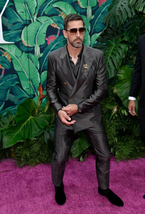 Aaron Rodgers in a double-breasted suit on a red carpet with tropical background. 