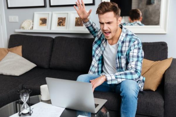 Photo of angry bristle man dressed in shirt in a cage print sitting on sofa in home and using laptop computer