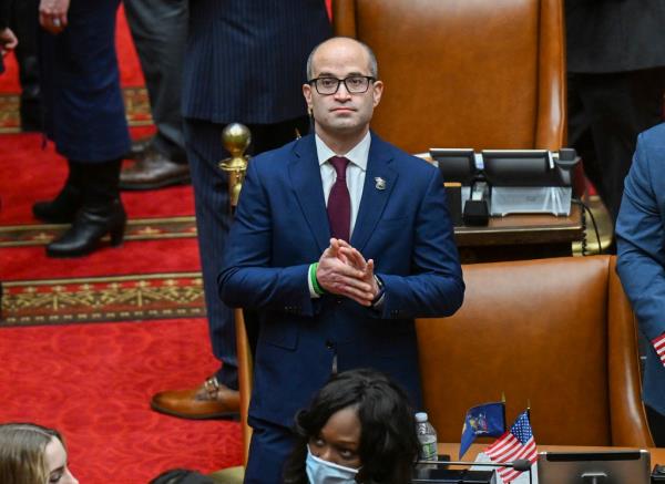 Ed Ra in a blue suit standing in the Assembly chamber next to a bunch of seats