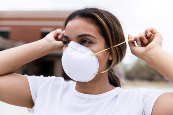 A mid adult woman protects herself by placing an N95 face mask over her nose and mouth.