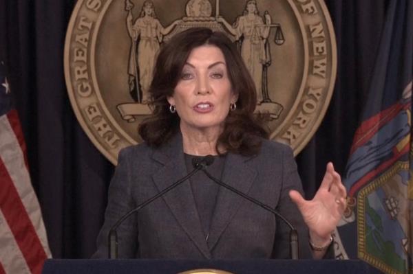 Hochul speaking at a podium with a microphone with a hand gesturing in front of the state seal