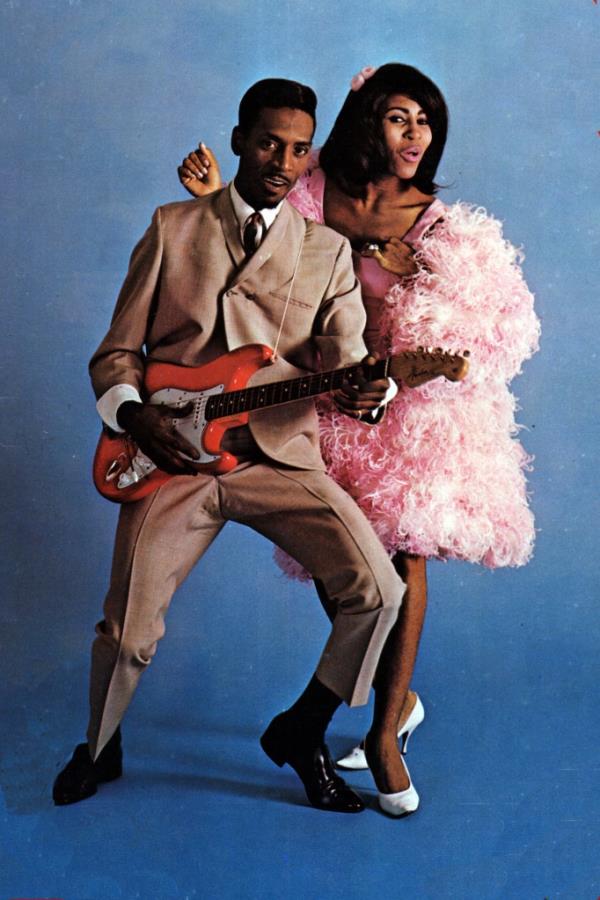 Ike and Tina Turner posing for portrait on blue background. Ike holds a red electric guitar while wearing a tan suit, while Tina is dressed in a pink, frilly frock