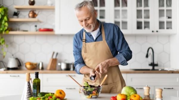 Happy senior man preparing healthy vegetable salad in kitchen, seaso<em></em>ning meal, smiling elderly gentleman in apron adding salt to bowl, enjoying cooking vegetarian food at home, free space