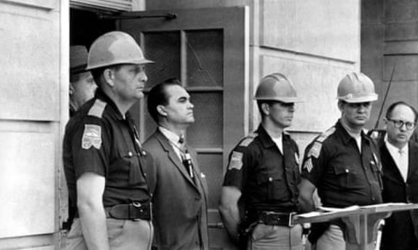 Black-and-white image of white man in suit flanked by white men in hard hats/riot hats in the doorway to a stone building.