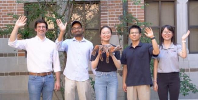 Five people smiling at the camera. Four are waving, while the person in the middle is holding a very large spider