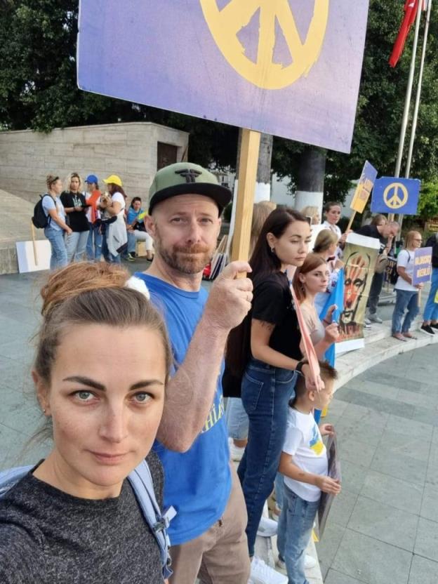 a white man and woman are seen at a rally. the man is holding a sign with a peace symbol painted on it