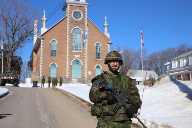 A man in military uniform smiles at the camera. he is carrying a gun and standing in front of a church. 