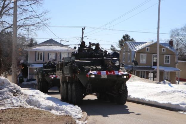 Two light armoured vehicles drive past residential homes. 