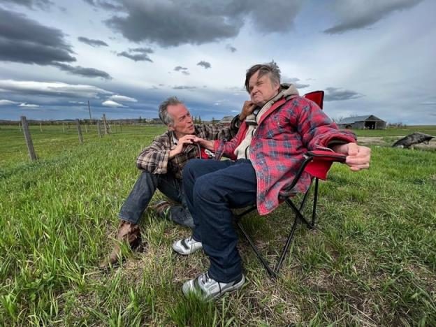 Two brothers, wearing plaid, are seen in a field of green grass. One man is sitting in a chair. The other is crouching beside him. 