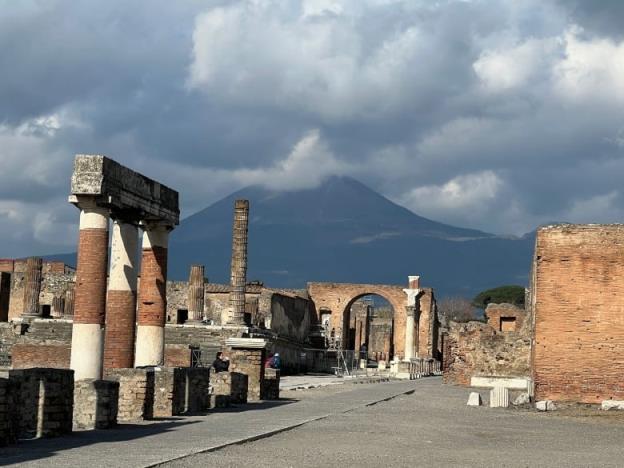 A streetscape of an ancient city with Mount Vesuvius volcano in the distance under a cloudy sky. 