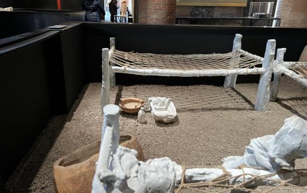 A museum exhibit shows bed f<em></em>rames with a rope net for lying on, along with pottery and other artifacts scattered on a pebbled floor. 