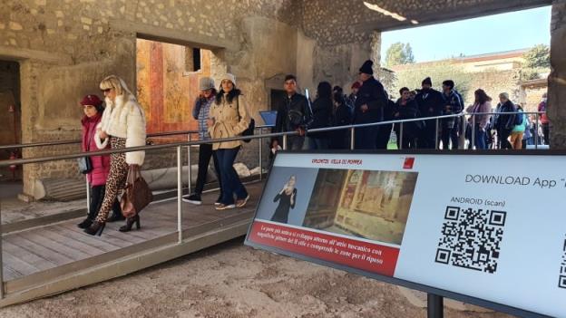 People walking through an archaeological museum pass by a mo<em></em>nitor that includes a sign-language interpreter as well as a QR code and some text in Italian.