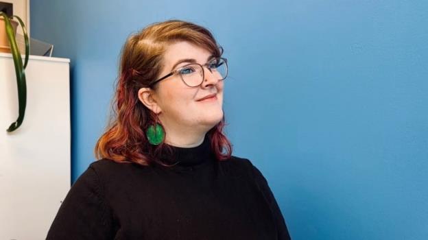 A woman with red hair, wearing glasses and large green earrings, stands in front of a bright blue wall.