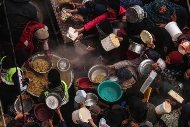 Women holding plates, bowls and buckets crowd together in front of people distributing food from a large me<em></em>tal pot. 