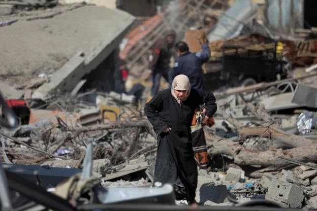 A woman wearing a head covering and long coat walks over co<em></em>ncrete rubble and debris.