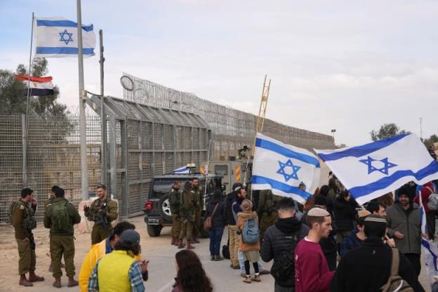 Protesters, some holding white and blue Israeli flags, stand in a crowd near a border fence, with uniformed soldiers and a military vehicle nearby.