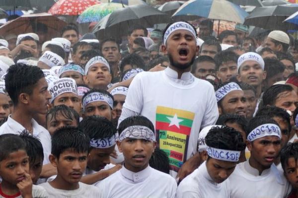 Young Rohingya men at a protest. They are dressed in white and have bandanas tied around their heads reading 'Genocide' One is wearing a T-shirt with the Myanmar flag. It is raining.