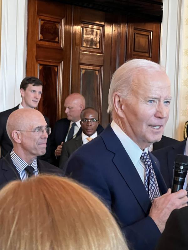 Joe Biden speaking into a microphone with Jeffrey Katzenberg behind him, in a White House room.