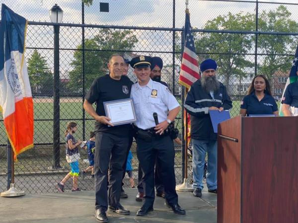 Esposito at a past event in Queens outdoors in park, holding a certificate being given by officer with other attendees in background.