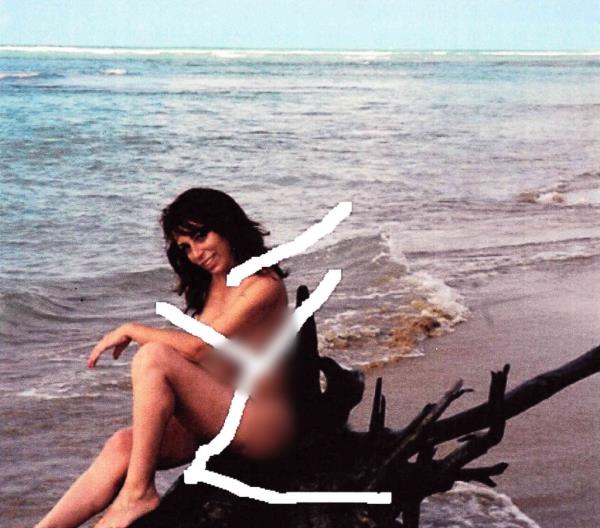 Cecilia Reynolds posing naked while sitting on driftwood on a beach at the tideline.