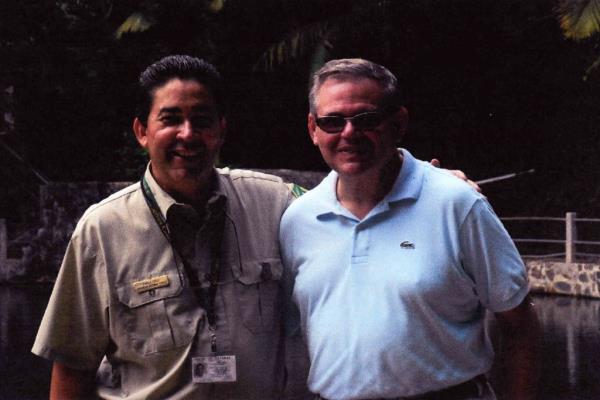 Bob Menendez in Lacoste tennis shirt posing in front of water and palm trees with man in uniform of the United States Forest Service.