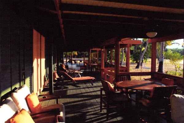 A shaded porch with sun loungers and a table with four chairs. Palm trees in the background.