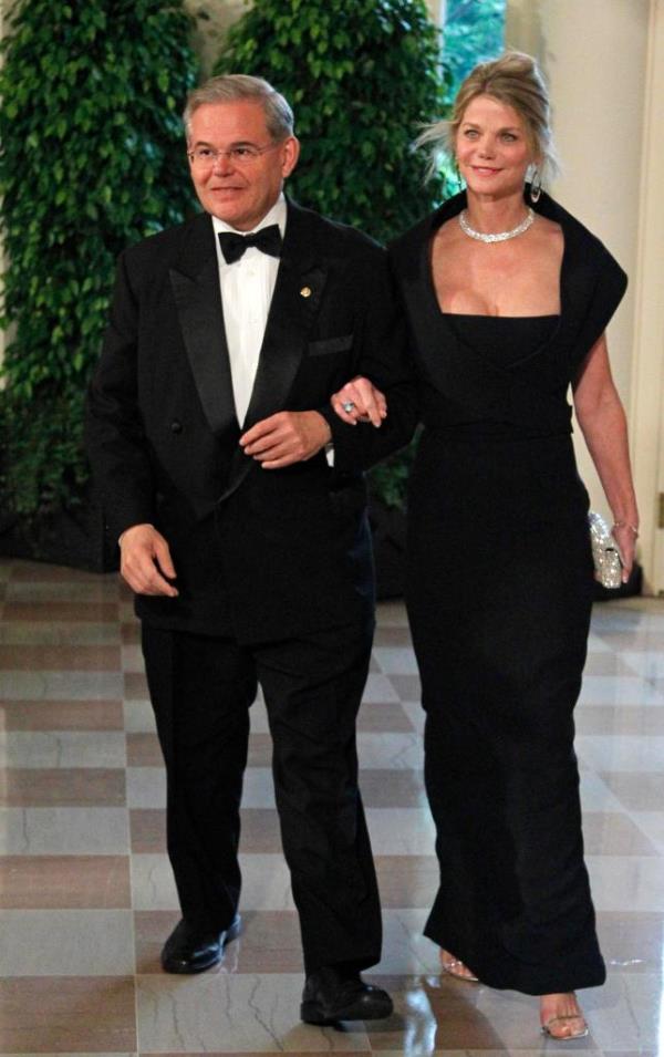 Bob Menendez in a tuxedo and black bow tie with Gwendolyn Beck in a black floor-length ballgown. She has her arm through his. They are walking on a checker-tiled floor in front of green foliage.