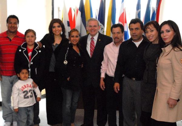 A group showing Bob Menendez in the center in front of flafs with Cecilia Reynolds at the far right and a group of people smiling including a child in a Jordan 23 top.