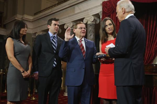 Bob Menendez raising his right hand, with his left hand on a bible, facing Joe Biden. His daughter in a black sleeveless dress is holding the bible and two people are standing behind him: his son Robert Jr. and a woman in a black sleeveless dress.