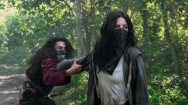 Stephanie Beatriz wearing a bandanna covering her face below the eyes, standing outside. 