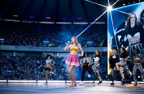 Taylor Swift, dressed in a short pink skirt and gold halter top, performs to a big crowd, with backup dancers clad in black and white outfits and a closeup screen behind her.