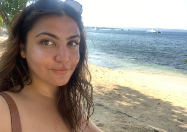 Almara Abgarian taking a selfie on a beach, wearing sunglasses (head and shoulders visible), with sea and sand in the background.