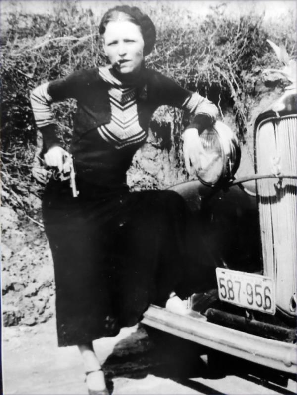 Bo<em></em>nnie with a cigarette in her mouth, a revolver in her hand, which is on her hip, leaning on the front headlight of her and Clyde's getaway Ford V8.