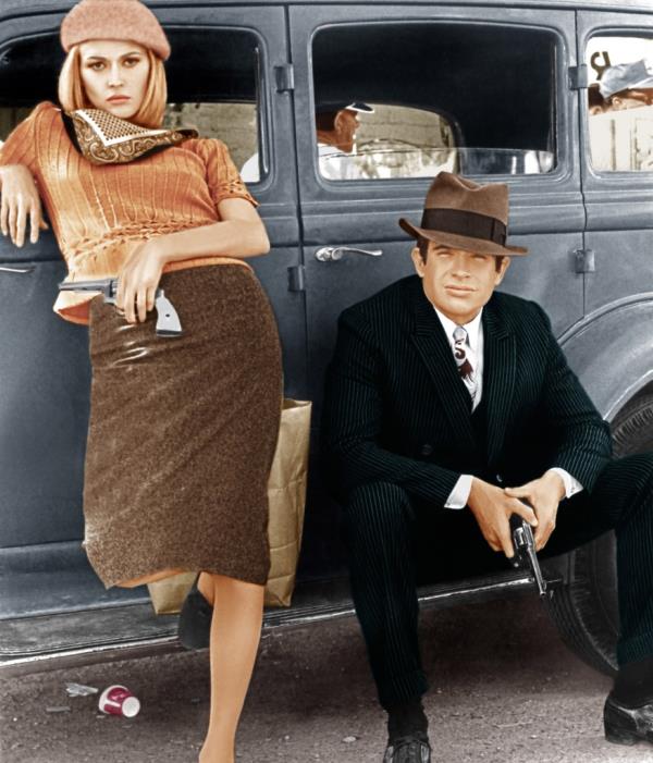 Faye Dunaway, revolver in hand, standing with right elbow on driver-door of car, Warren Beatty sitting on running board also carrying revolver, in publicity still from 