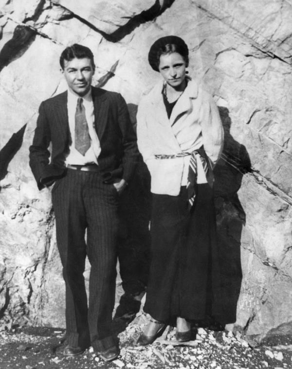 Bo<em></em>nnie and Clyde posing in front of a sheer rock face. Clyde, wearing a suit with open jacket, white shirt and tie has his hands in his pockets. Bonnie, in light jacket tied apparently by a scarf or necktie, has wide-legged pants or a long skirt, heels and beret, and stands with hands behind back, 