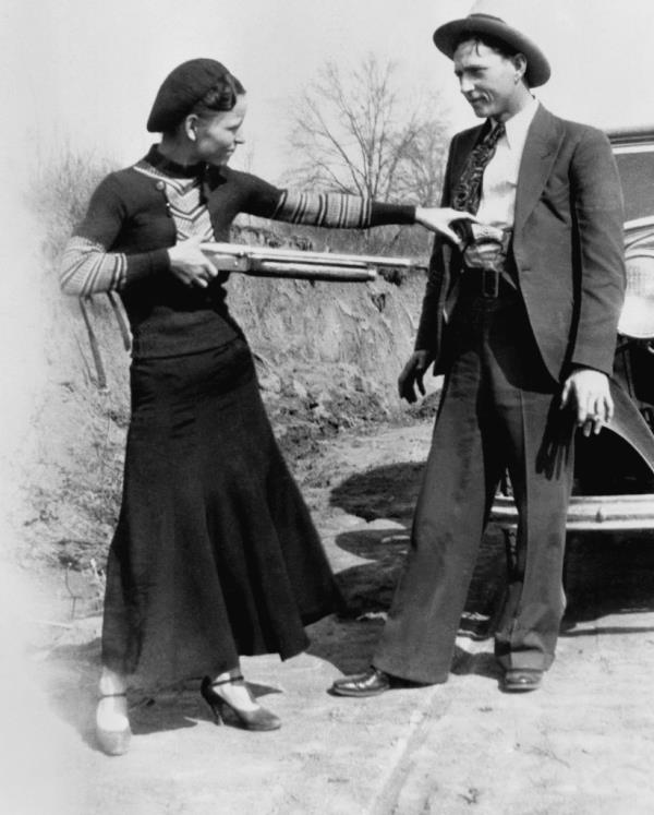 Bo<em></em>nnie holds a shotgun on her right hand apparently aimed at Clyde's stomach. She has her left had on the handle of a revolver tucked in his belt. He is wearing an open suit, white shirt and tie and they stand in front of their V8.