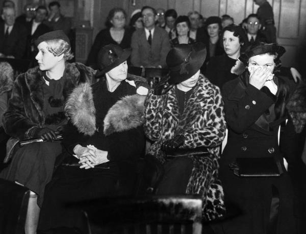 Four women, two in fur-lined coats, one in apparent animal-patterned fur, and one in black, all wearing 1930s-style hats. One has a handkerchief over her lower face. 