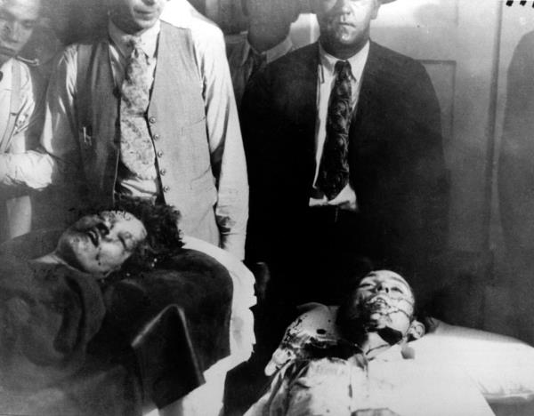 Two men, one in a suit and tie, the other light shirt, open vest and tie, stand behind the bloodied corpses of Bo<em></em>nnie and CLyde on gurnies.