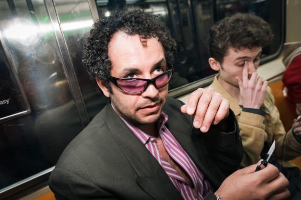 Kareem Rahma sitting on the subway, wearing purple glasses and looking up. 