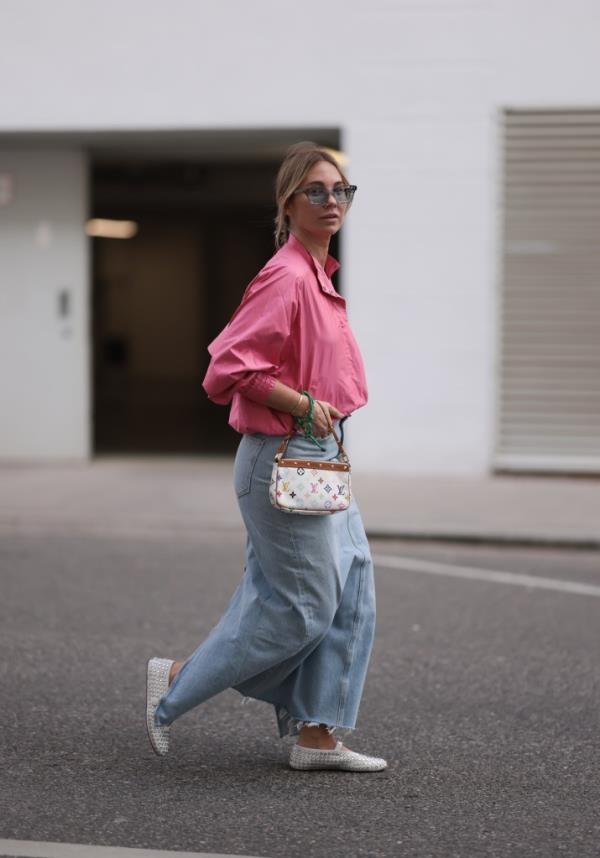 Karin Teigl wearing grey sunglasses, pink vintage jacket, light blue denim skirt, Louis Vuitton white canvas bag with colorful logo, green bracelet, and white rhinestone-patterned ballerinas.